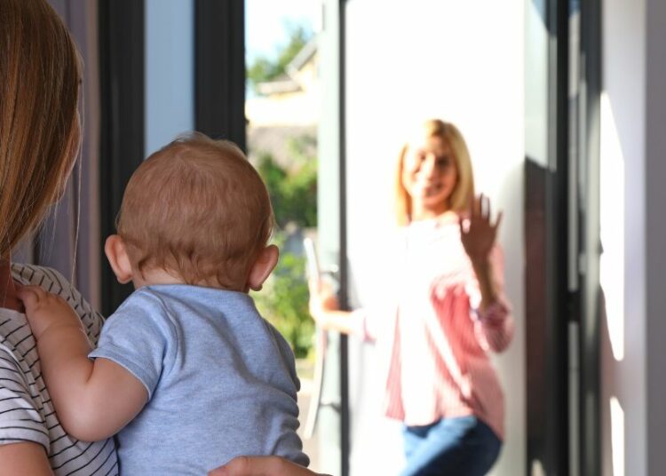 une femme sort par la porte d'entree en saluant une jeune femme qui porte un bebe dans ses bras