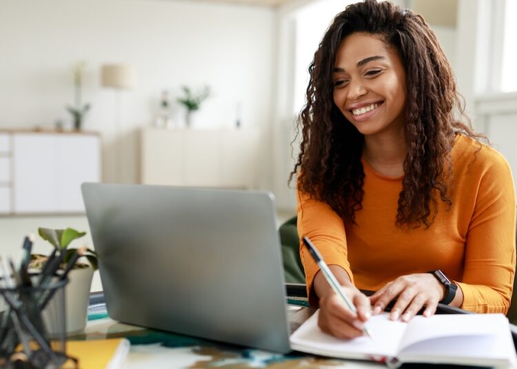 Smiling young African American woman sitting at desk working on laptop taking notes in notebook, happy millennial female studying online, watching webinar using computer and writing check list