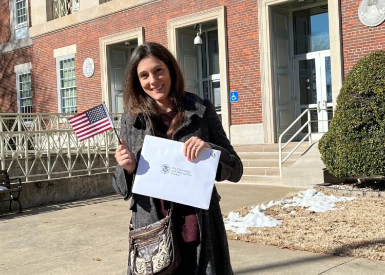 Claire Speaks se tient devant un bâtiment de l'administration américaine avec un drapeau américain et son certificat de citoyenneté.